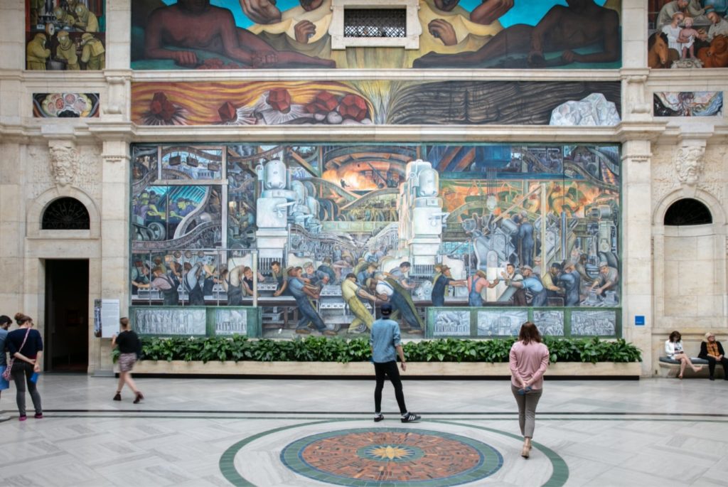 Several people look at Diego Rivera's mural at the DIA, a multi-story image depicting Detroit's workers.