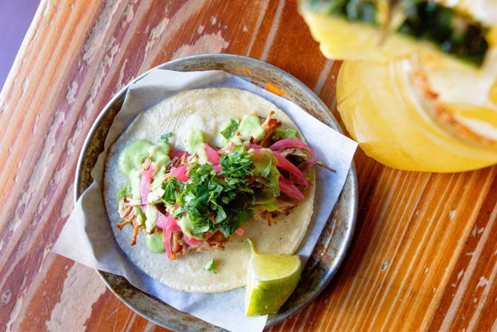 A taco on a wooden table, topped with pickled red onions and cilantro.
