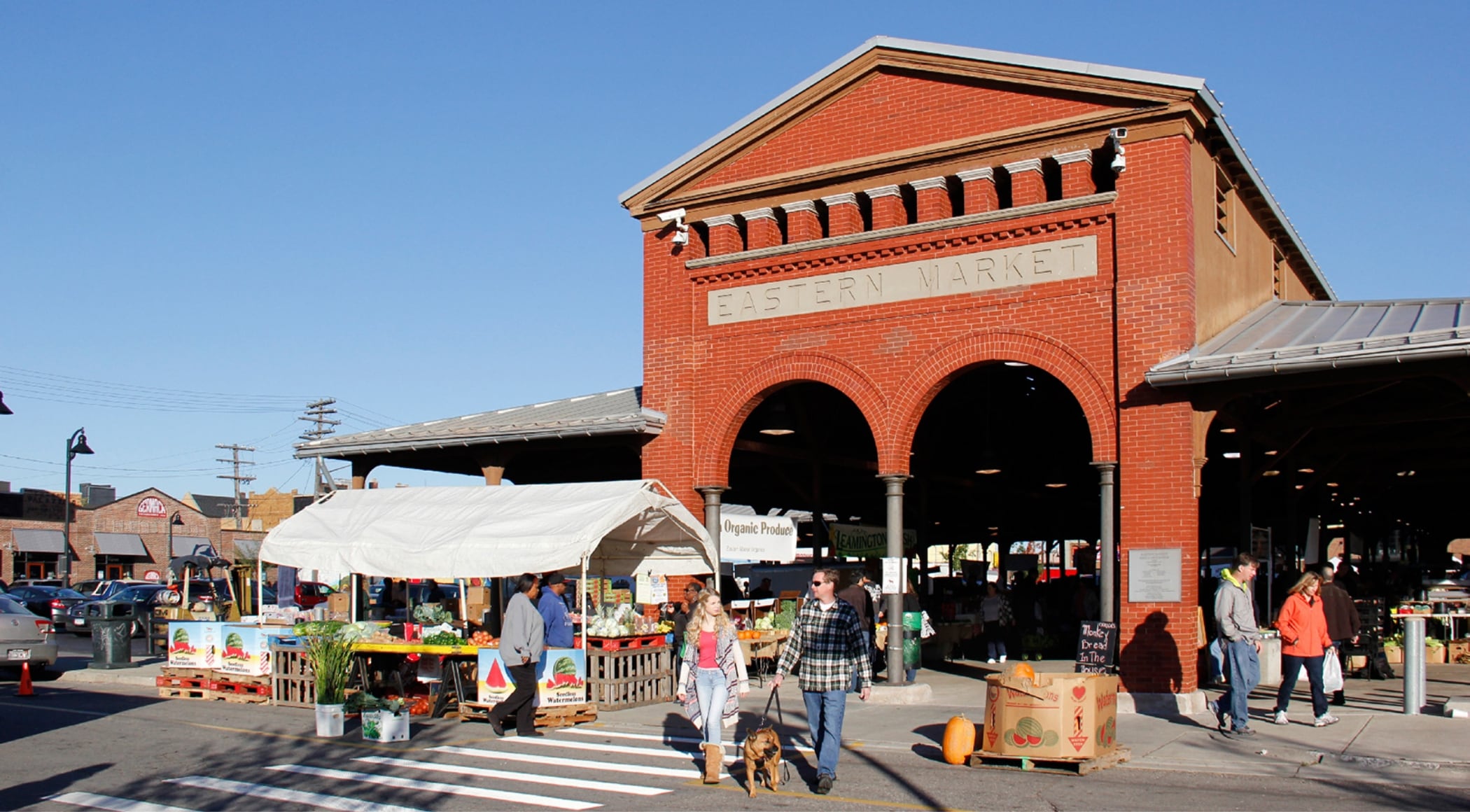 Easter Market's iconic red brick shed with arched entryway. In front of it sits a white tent full of vegetables and several people browsing.
