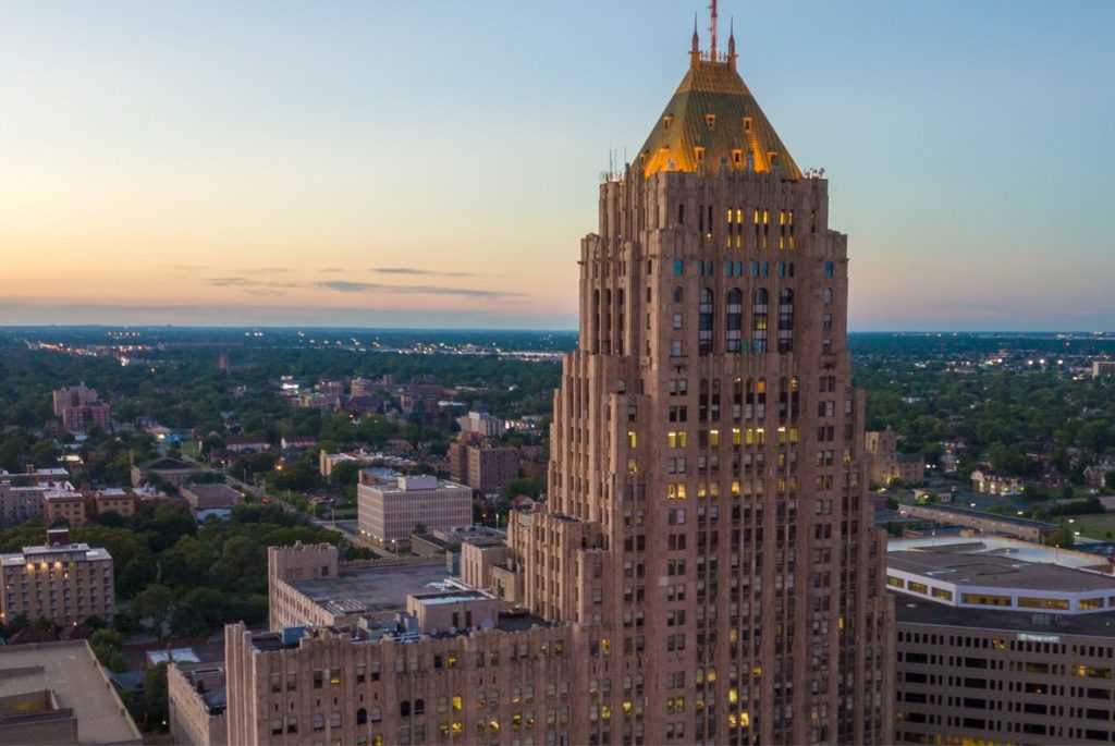 The Fisher Building shines above Detroit, showcasing stoneworks and its iconic turquoise roof. Behind the building is an expanse of trees and shorter buildings at sunset.