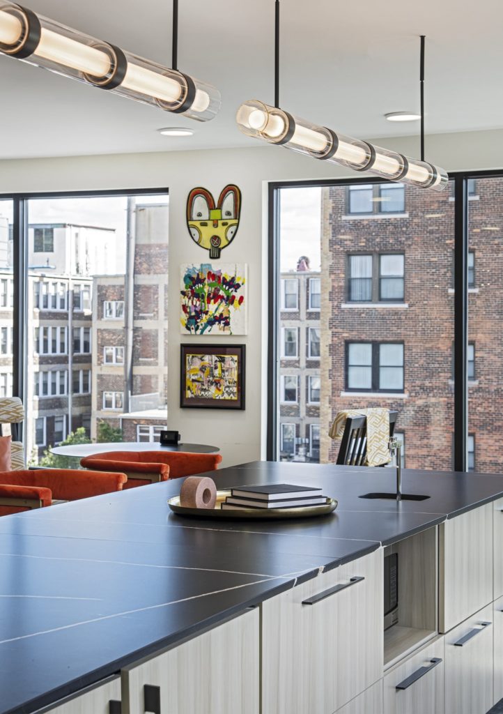 A island with a black countertop and soft orange stools. Behind are large windows with views of Detroit.