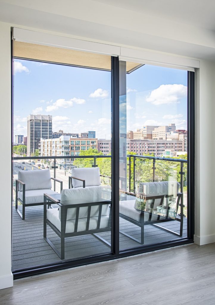 A corner balcony is visible behind large sliding glass doors. Four cushioned chairs overlook skyline views of Detroit.